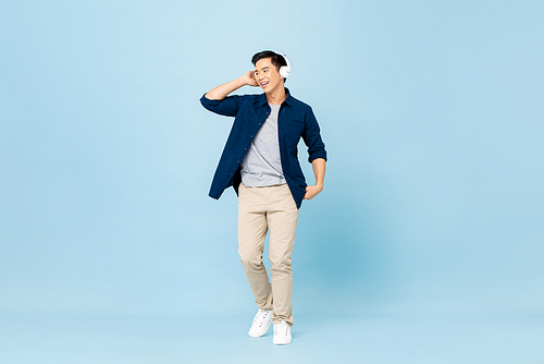 Full body portrait of smiling young handsome Asian man listening to music with wireless headphones isolated on light blue studio background