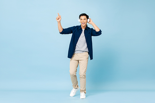 Full body portrait of happy smiling young handsome Asian man listening to music with wireless headphones pointing hand up on isolated light blue studio background