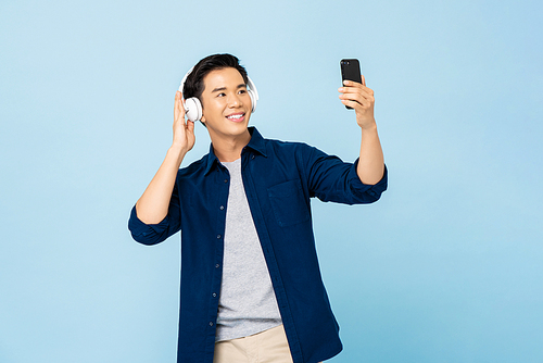 Happy smiling Asian man wearing headphones listening to music from mobile phone in light blue isolated studio background