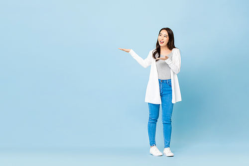 Young cheerful beautiful Asian woman with hands opening to copy space aside on isolated light blue studio background