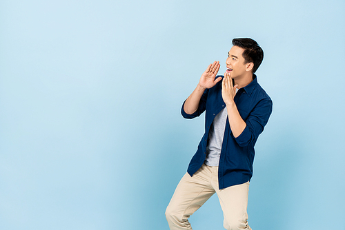 Young Asian man shouting and cupping hands around mouth in light blue isolated background with copy space