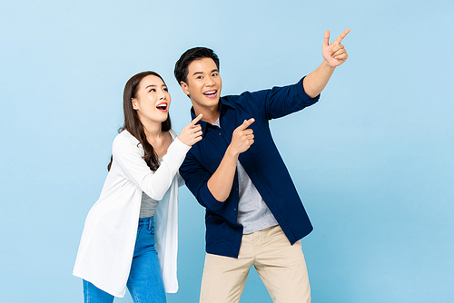 Smiling excited Asian couple pointing hands upward to empty space on isolated light blue background