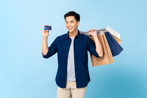 Happy smiling handsome Asian man carrying shopping bags and showing credit card in light blue isolated studio background