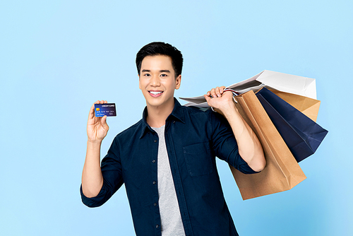 Handsome Asian man carrying shopping bags showing credit card in hand isolated on light blue background