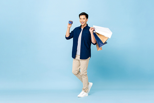 Full lenght portrait of happy smiling handsome Asian man carrying shopping bags and showing credit card in light blue isolated studio background