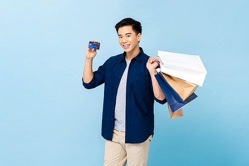 Happy smiling handsome Asian man carrying shopping bags and showing credit card in light blue isolated studio background