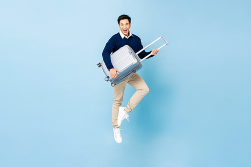 Portrait of young energized Asian tourist man with baggage jumping in mid-air ready to travel isolated on light blue studio background