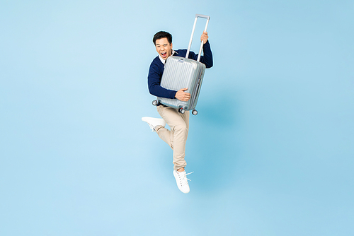Portrait of young excited handsome Asian tourist man with baggage jumping in mid-air ready to travel isolated on light blue studio background