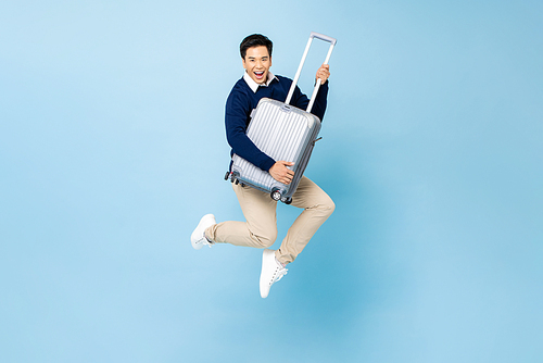 Fit healthy smiling young handsome Asian tourist man with baggage jumping in mid-air ready to fly isolated on light blue studio background