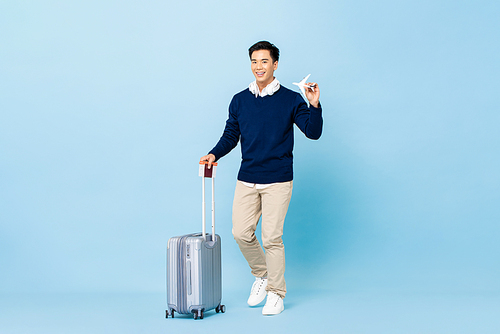 Young smiling handsome Asian tourist man with baggage, passport and boarding pass ready to fly on light blue isolated studio background