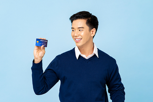 Happy smiling young handsome Asian man holding credit card in isolated light blue background