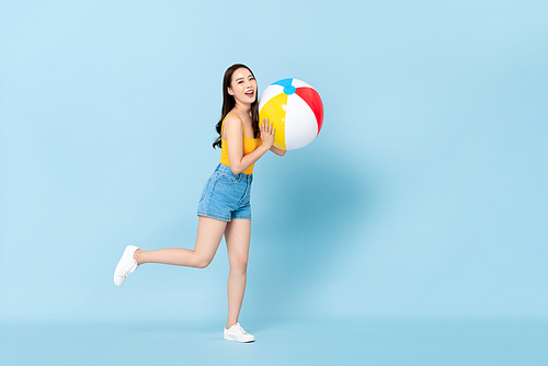 Smiling pretty Asian girl in summer outfit with colorful beach ball on isolated light blue background