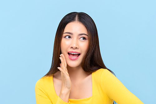 Young pretty Asian woman saying something with hand beside mouth on light blue isolated background
