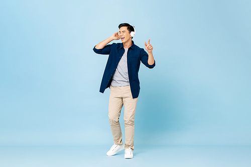 Smiling enjoyful young handsome Asian man listening to streaming music on wireless headphones isolated on light blue studio portrait background