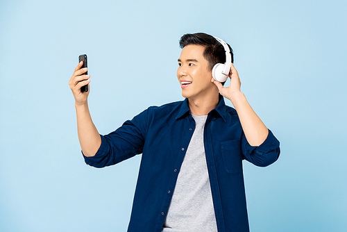 Happy smiling Asian man wearing headphones listening to music from mobile phone in light blue isolated studio background