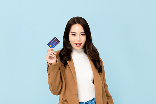 Beautiful smiling Asian woman in winter clothes showing credit card in hand for financial and cashless society concepts isolated on light blue  background