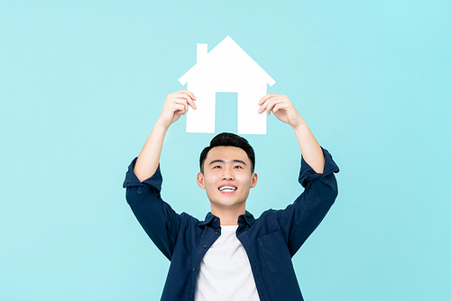 Happy young Asian man holding house sign overhead isolated on light blue background for real estate concepts