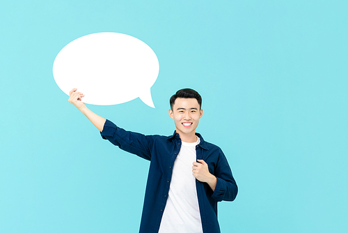 Waist up portrait of young smiling Asian man holding up an empty white speech bubble in light blue studio background