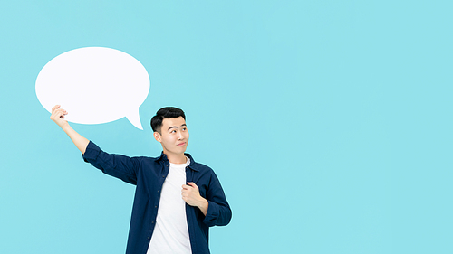 Young smiling Asian man holding empty white speech bubble whlie thinking and looking at copy space in light blue studio background