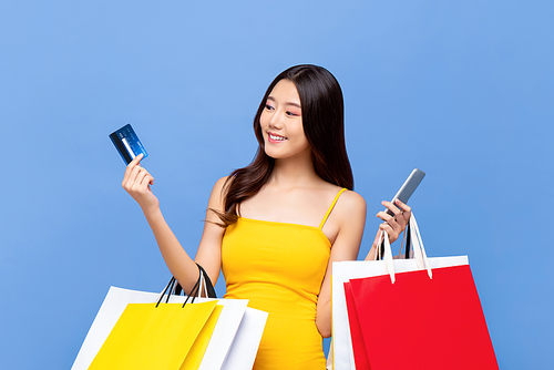 Young smiling beautiful Asian woman making online payment through mobile phone via credit card while carrying colorful shopping bags in isolated blue background