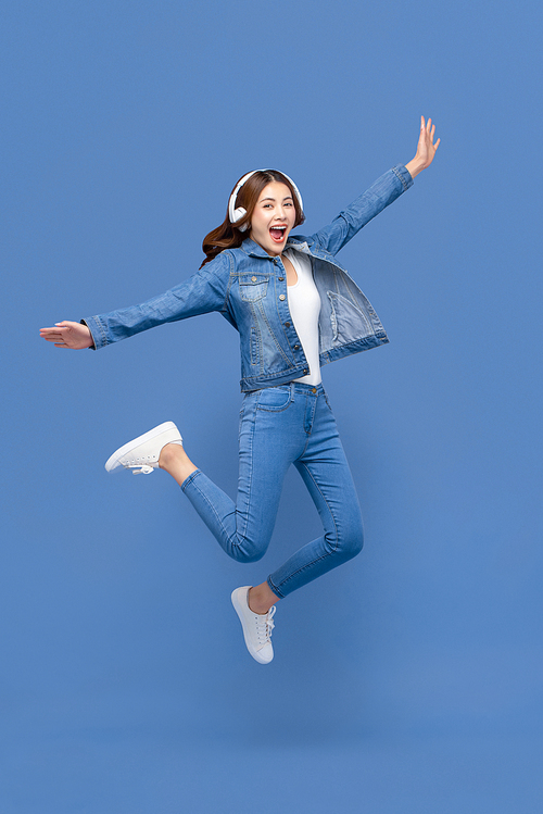 Happy young Asian woman jumping while listening to music on headphones isolated on blue studio background