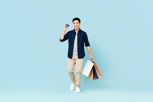 Handsome young Asian tourist man carrying bags shopping with credit card isolated on light blue background