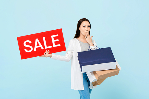 Surprised beautiful Asian woman with shopping bags showing red sale sign isolated on light blue background