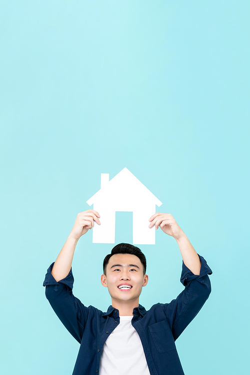 Happy young Asian man holding house sign with copy space  overhead isolated on light blue background for real estate concepts