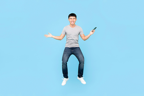 Fun full length portrait of happy smiling Asian man jumping in mid-air while holding smartphone in isolated studio blue background