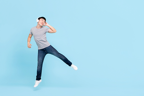 Smiling energetic young Asian man wearing headphones listening to music jumping and looking at copy space on light blue isolated background