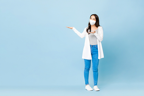 Young Asian woman wearing face mask opening hands to empty space aside in isolated light blue studio background