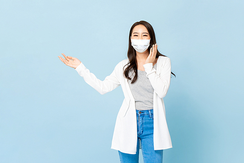 Young Asian woman wearing face mask opening hand to empty space aside in isolated light blue studio background
