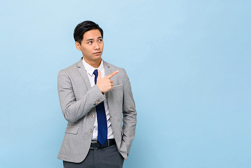 Young handsome Asian businessman in coporate wear pointing hand to empty space aside in isolated blue studio background