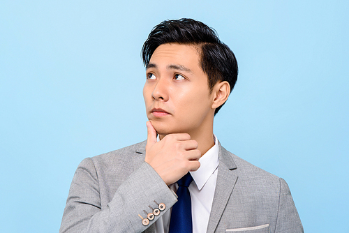 Close up portrait of serious young handsome Asian businessman thinking with hand on chin isolated studio blue background