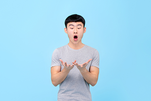 Fun portrait of astonished young Asian man looking at both hands with mouth open in isolated studio blue background