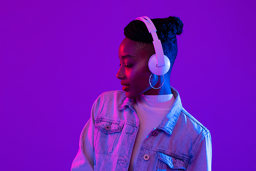 Young African American woman wearing headphones listening to music in futuristic purple cyberpunk neon light background