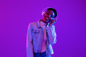 Happy young African American woman wearing headphones listening to music on  purple neon light background