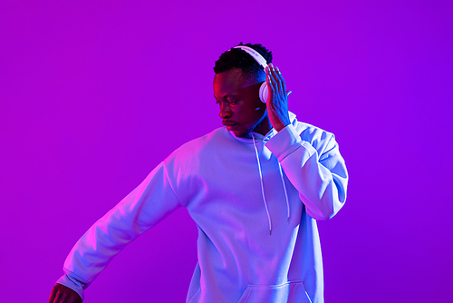 Young handsome African man wearing headphones listening to music and dancing in futuristic purple cyberpunk neon light background
