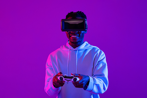 Young African man wearing VR glass headset playing game with joystick controller in futuristic purple cyberpunk neon light background