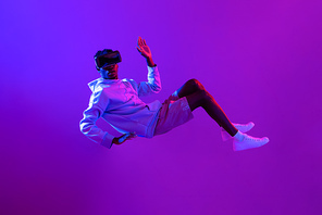 Amazed young African man wearing VR glass headset levitating in the air on futuristic purple cyberpunk neon light background