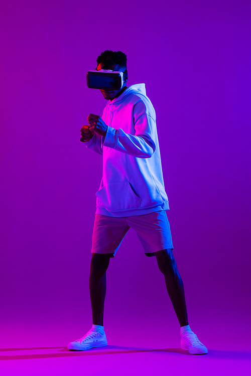 Full length portrait of young African man wearing VR glass headset standing and punching in futuristic dark purple cyberpunk neon light background