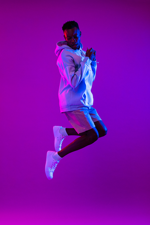 Full lenght portrait of young energetic African American man jumping in dark purple futuristic cyberpunk neon light background