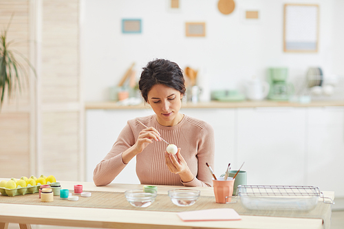 Portrait of modern adult woman painting Easter eggs while sitting at table in cozy kitchen interior, copy space