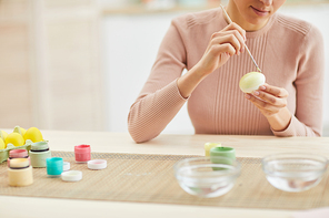 Cropped portrait of smiling adult woman painting Easter eggs while sitting at table in cozy kitchen interior, copy space