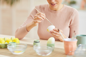 Cropped portrait of smiling young woman painting Easter eggs while sitting at table in cozy kitchen interior, copy space