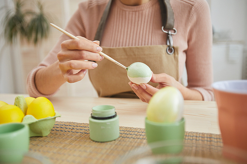 Cropped portrait of young woman painting Easter eggs while sitting at table in cozy kitchen interior, copy space
