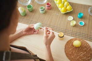 High angle close up of unrecognizable young woman painting eggs in pastel colors for Easter while sitting at table in kitchen or art studio, copy space