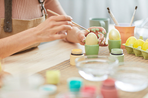 Close up of unrecognizable young woman painting eggs in pastel colors for Easter while sitting at table in kitchen or art studio, copy space