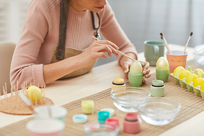 Cropped portrait of unrecognizable young woman painting eggs in pastel colors for Easter while sitting at table in kitchen or art studio, copy space