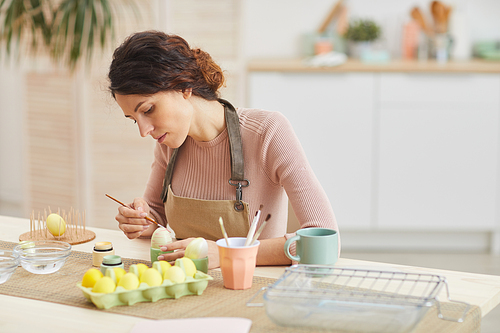 Portrait of elegant young woman painting eggs in pastel colors for Easter while sitting at table in cozy kitchen interior, copy space